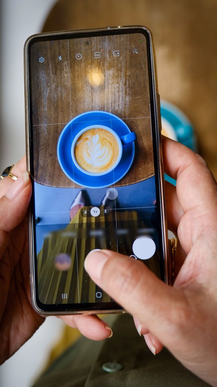 カフェの席で、スマートフォンを縦に構えてラテを撮っている手元